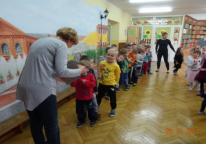 Dzieci stoją w dwóch rzędach. Nauczycielka trzyma serduszko, które dzieci przekuwają.